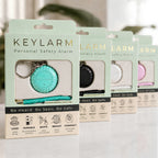 Four Keylarm personal safety alarm keychains in different colors (mint green, black, white, and pink) displayed in their retail packaging on a light marble surface.