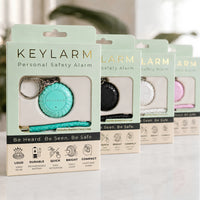 Four Keylarm personal safety alarm keychains in different colors (mint green, black, white, and pink) displayed in their retail packaging on a light marble surface.