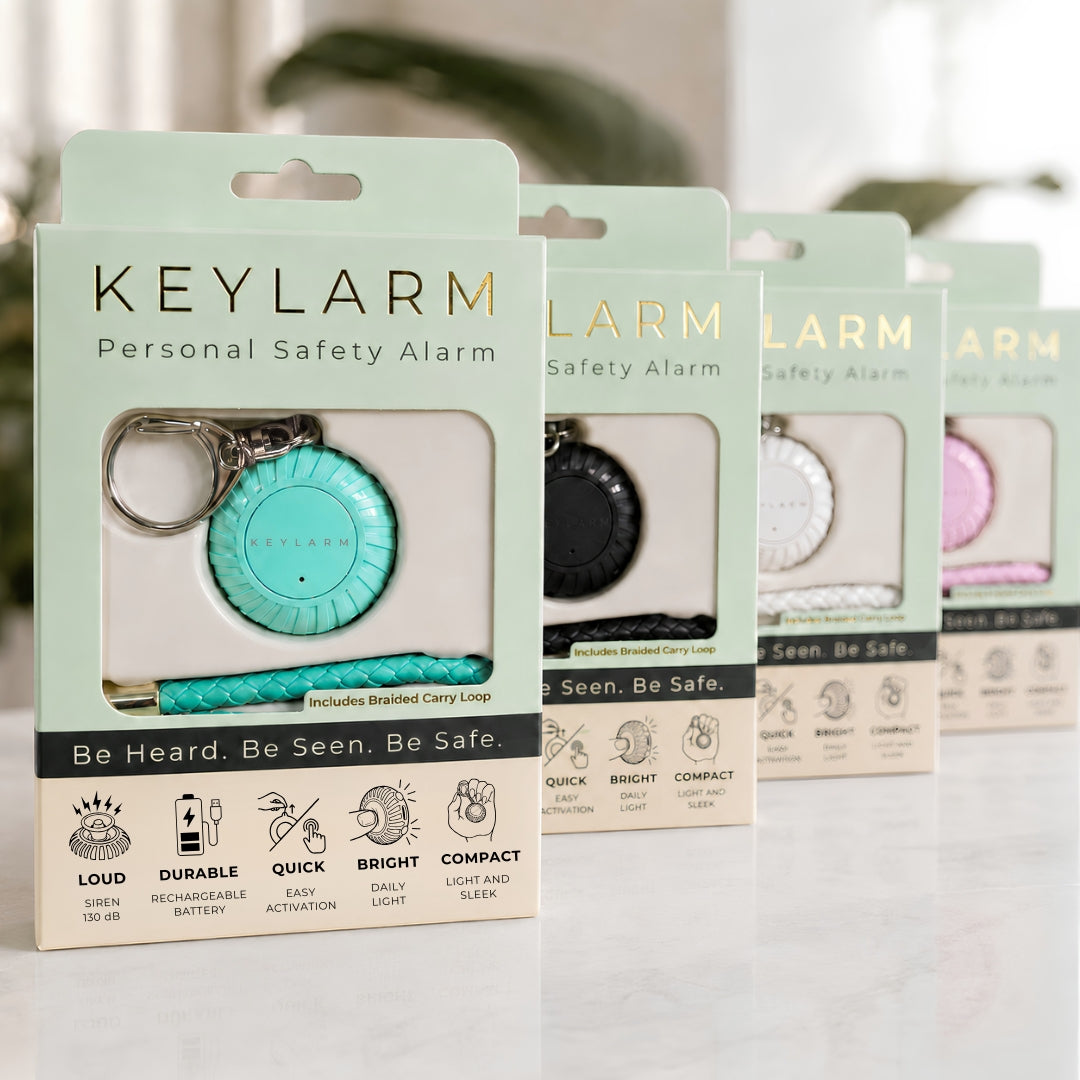 Four Keylarm personal safety alarm keychains in different colors (mint green, black, white, and pink) displayed in their retail packaging on a light marble surface.